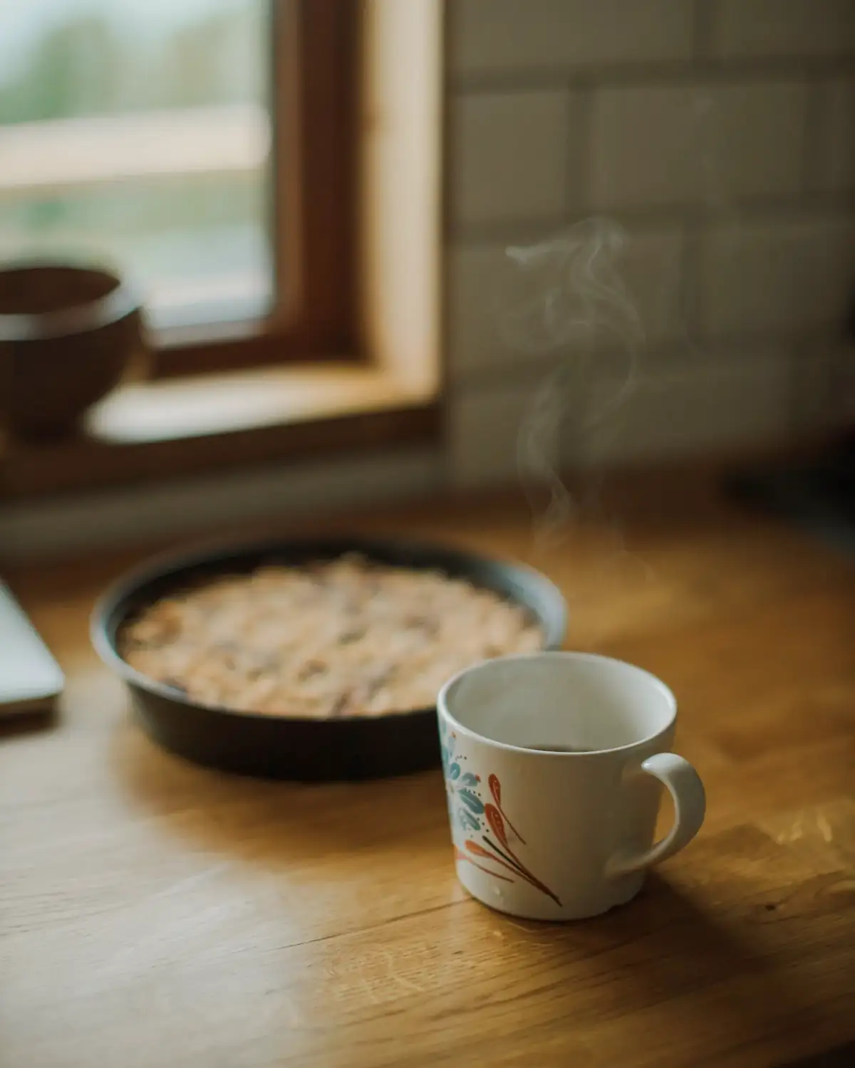 Een mok koffie naast een taart in de vorm, op een houten tafel bij het raam